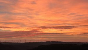 Durch Sonnenuntergang Rotgefärbter Himmel mit Wolken. Am unteren Bildrand die Landschaft mit vereinzelten