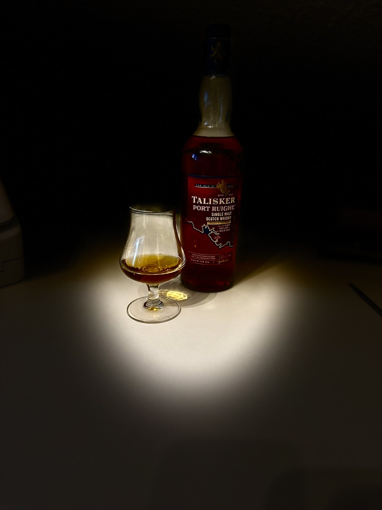 Eine Flasche Talisker Port Ruighe Single Malt Scotch Whisky steht auf einem Tisch, daneben ein halb gefülltes Nosing-Glas, beleuchtet von einem warmen Lichtkegel vor dunklem Hintergrund.