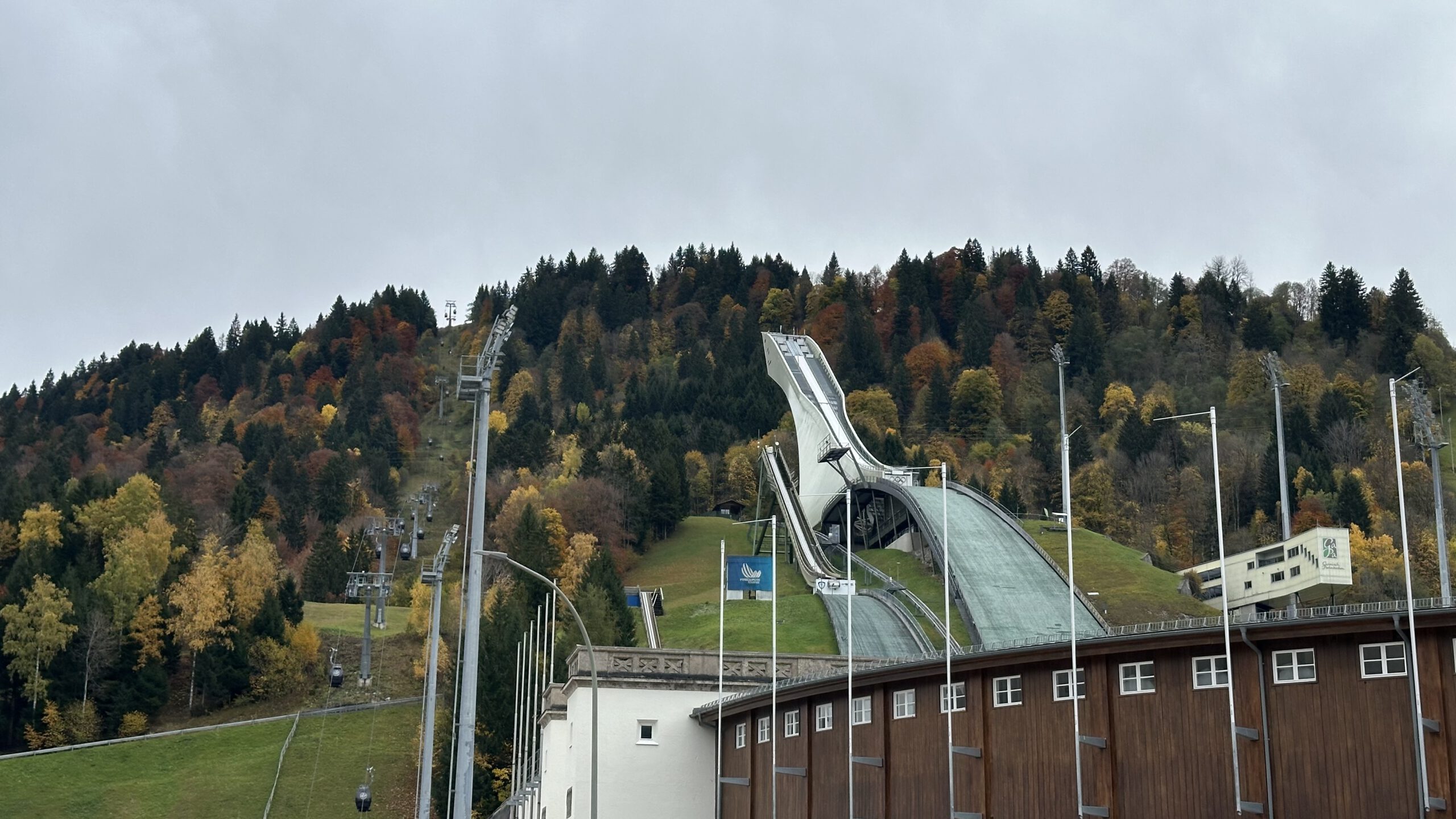 Blick auf die Große Olympiaschanze in Garmisch-Partenkirchen, umgeben von herbstlich gefärbtem Wald und bewölktem Himmel.