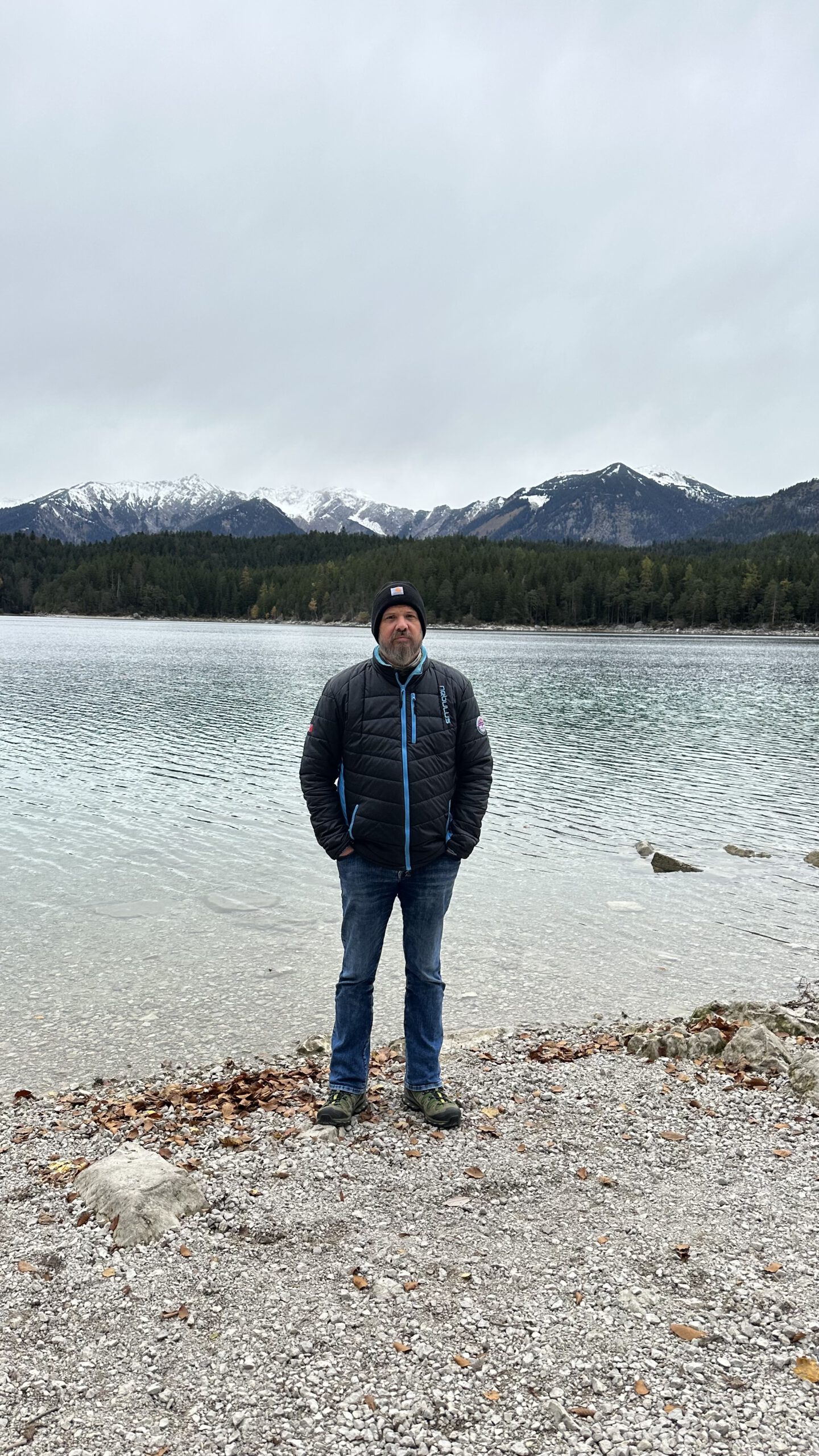 Ein Mann steht am Ufer des Eibsees bei Garmisch-Partenkirchen, im Hintergrund schneebedeckte Alpen und ein grauer Himmel.
