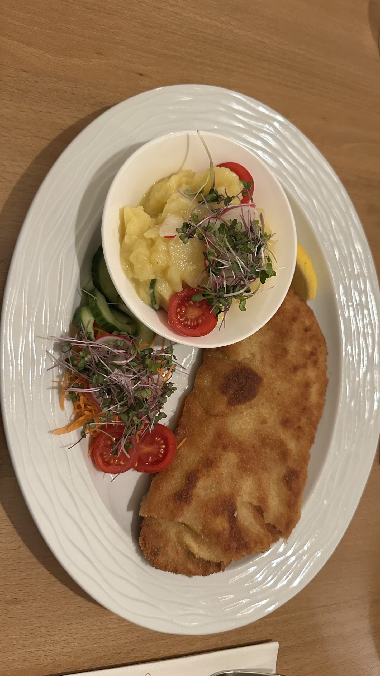 Goldbraun paniertes Schnitzel auf weißem Teller mit Kartoffel-Gurkensalat, Kirschtomaten und frischen Sprossen.