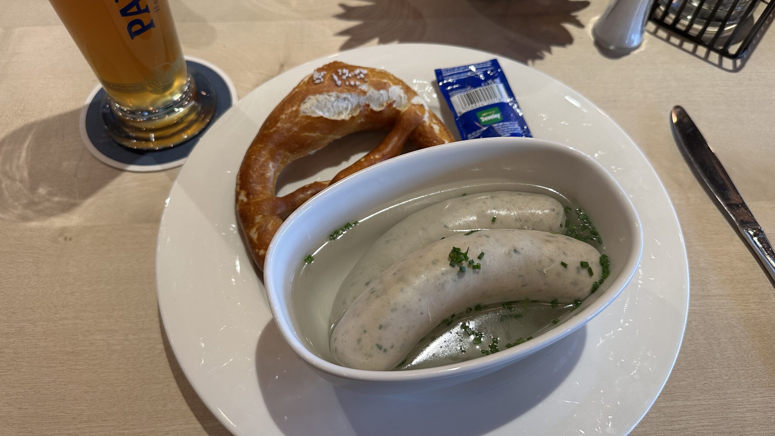 Zwei Weißwürste in heißem Wasser, serviert mit Brezel, süßem Senf und Weißbier auf einem hellen Holztisch.