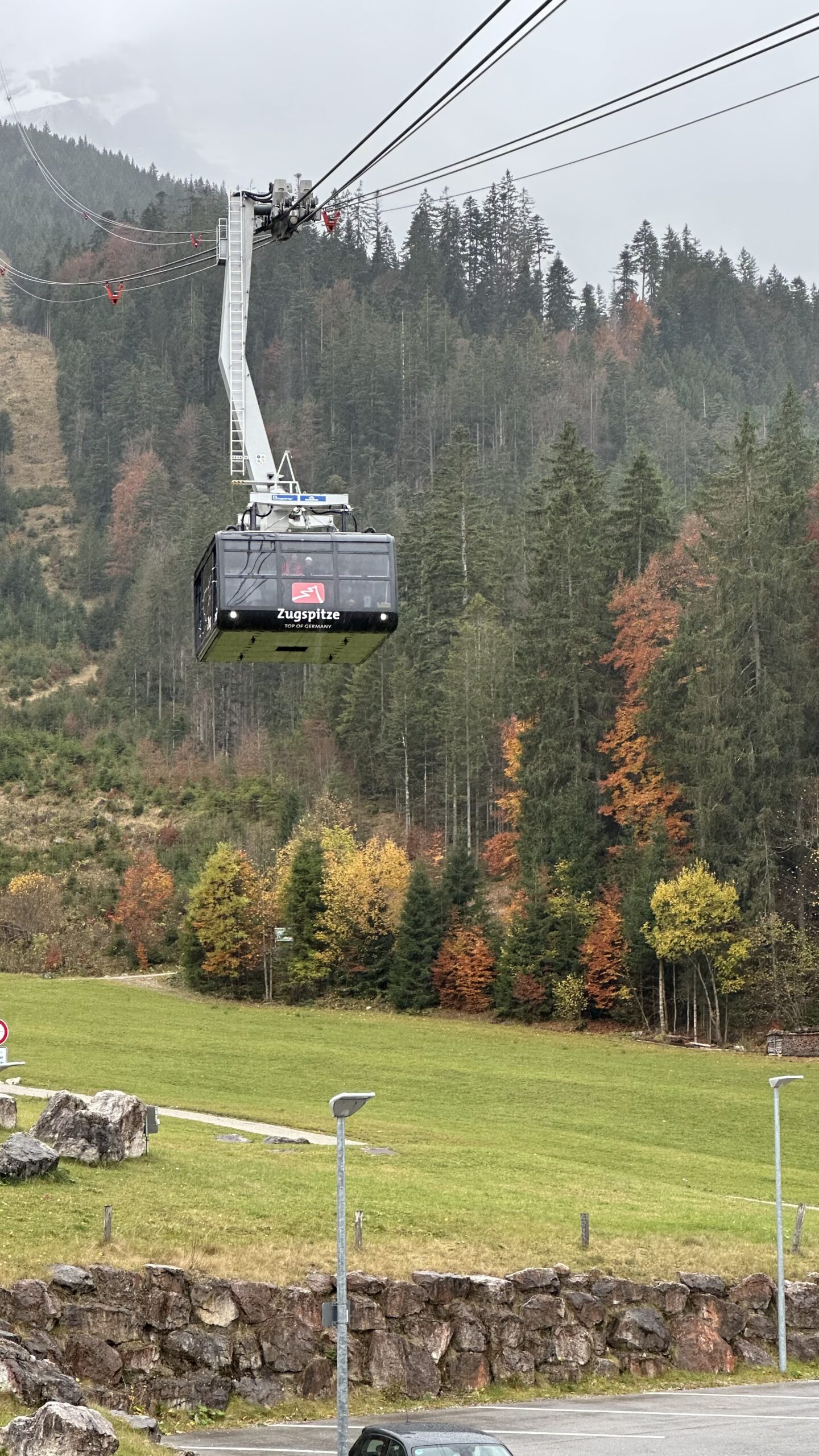 Gondel der Zugspitz-Seilbahn schwebt über einer grünen Alm mit herbstlich gefärbten Bäumen im Hintergrund.
