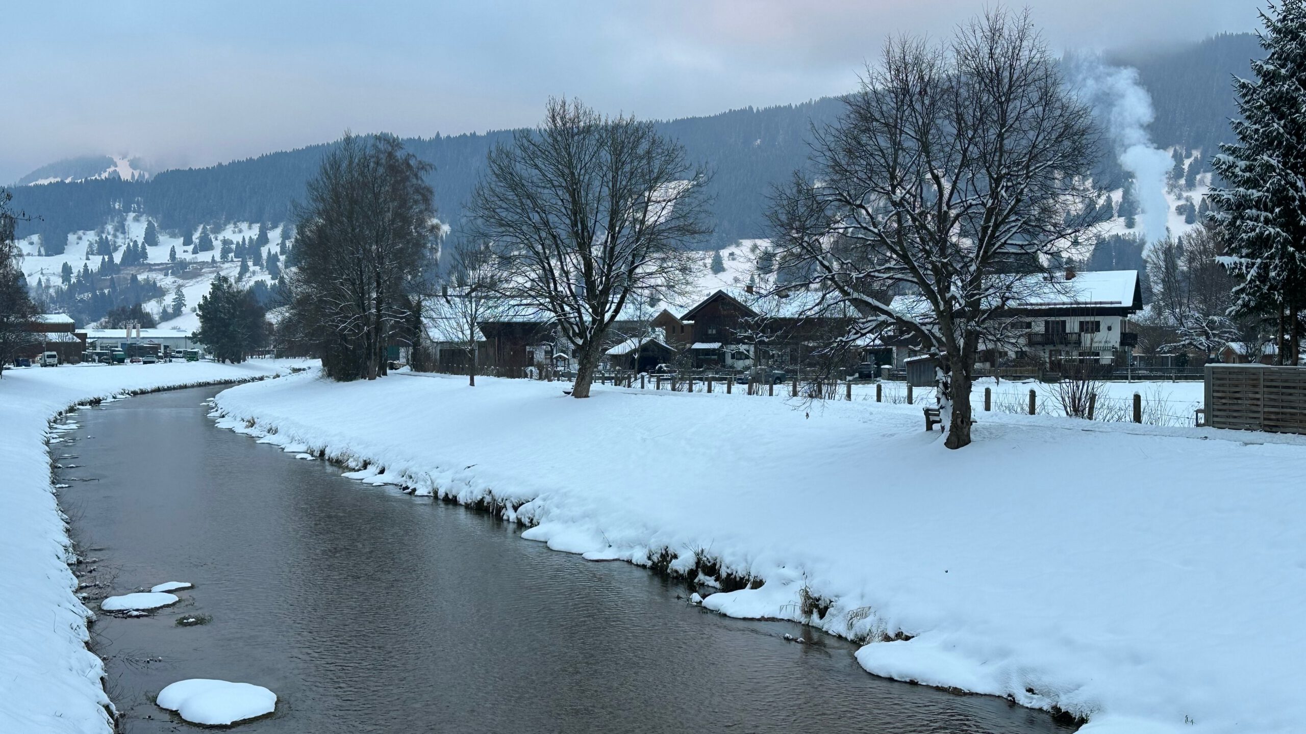 Winterlandschaft an der Ammer in Oberammergau mit schneebedeckten Ufern, Bäumen und Berghintergrund an einem nebligen Morgen.