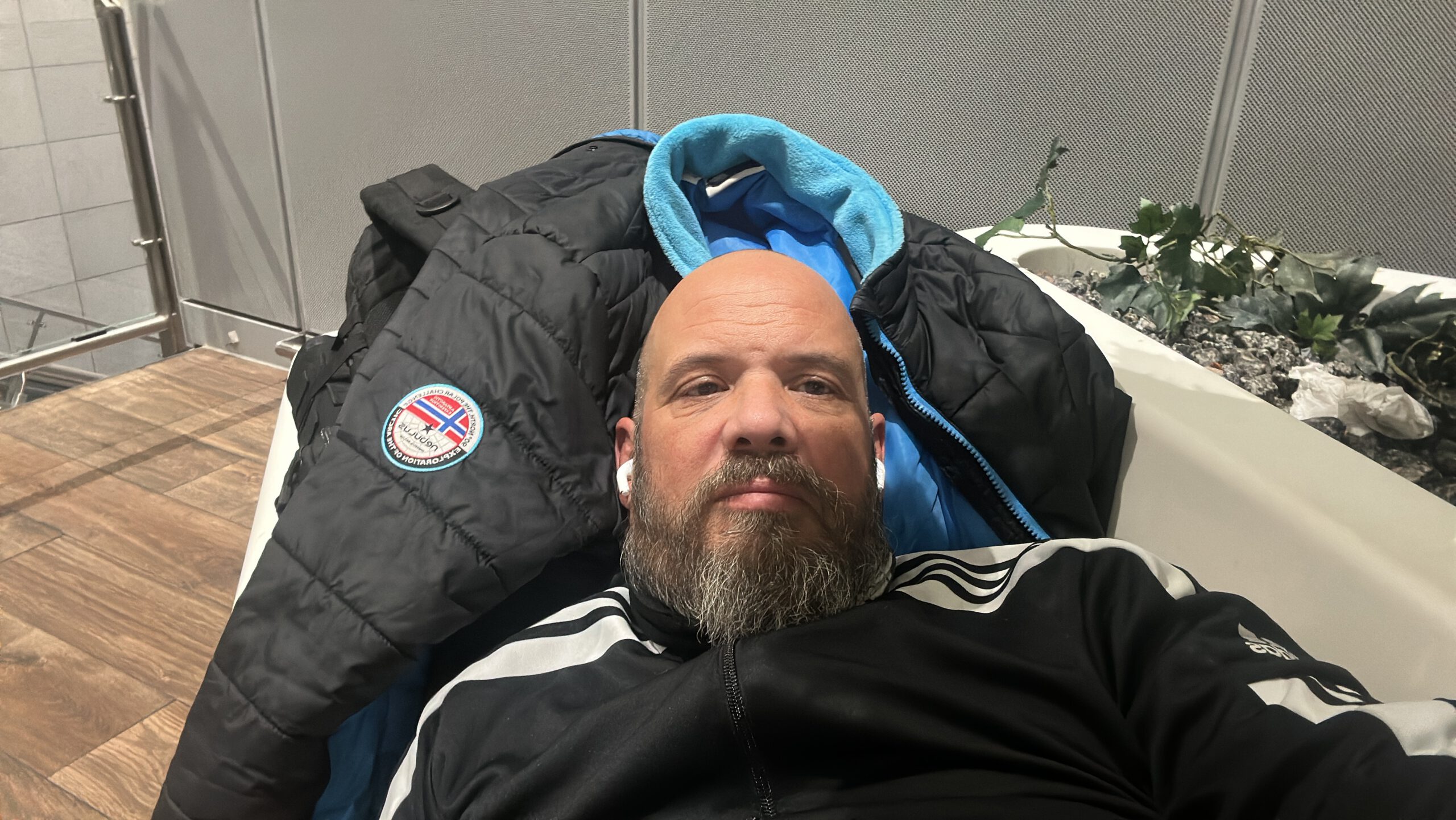 „Mann liegt nachts im Terminal 2 des Frankfurter Flughafens auf einer Bank, mit Jacke als Unterlage, umgeben von künstlichen Pflanzen und sterilem Flughafeninterieur.