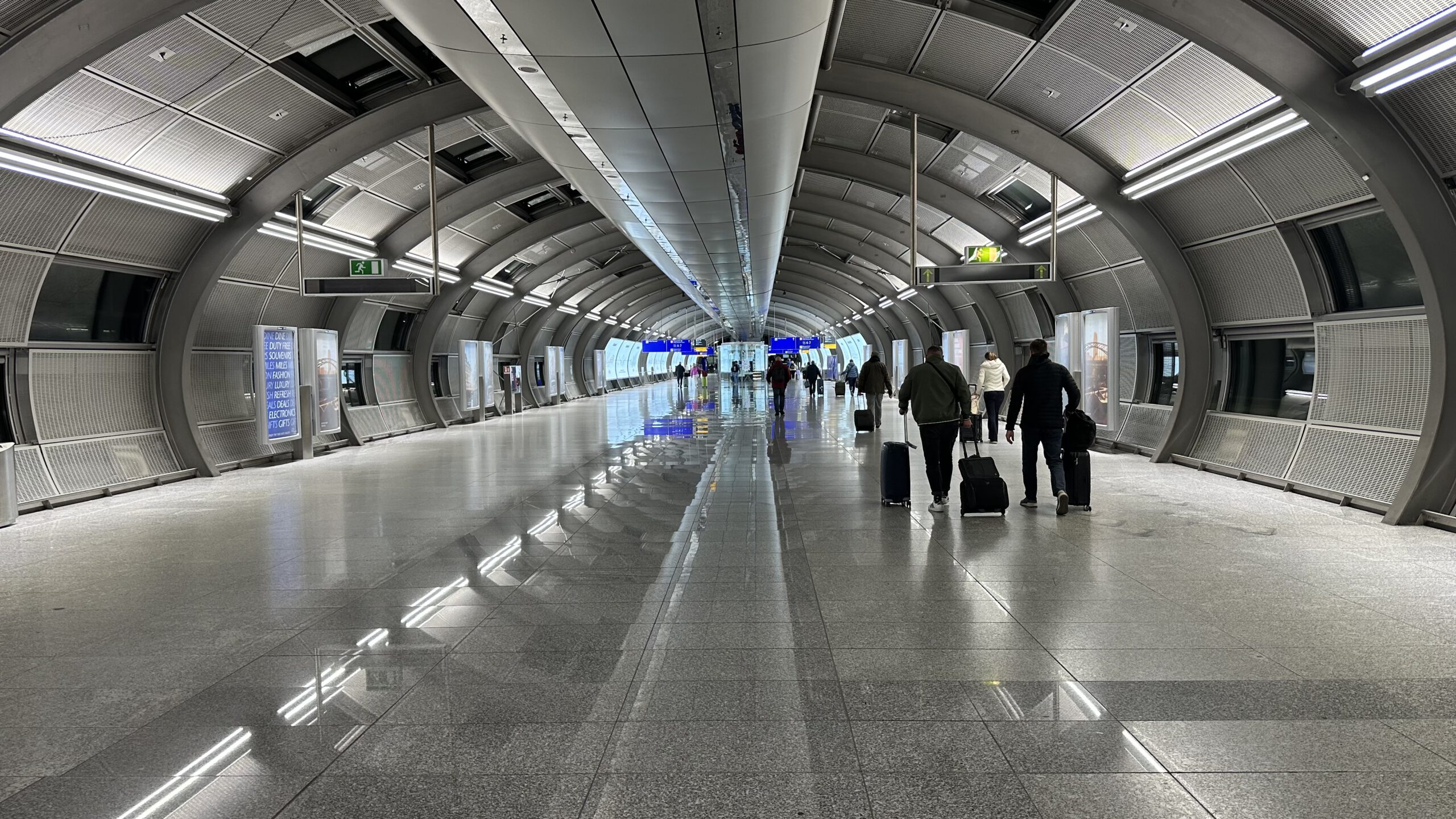 Futuristischer Passagiertunnel im Flughafen Frankfurt mit einigen Reisenden und rollenden Koffern, beleuchtet von kaltem, modernem Licht.
