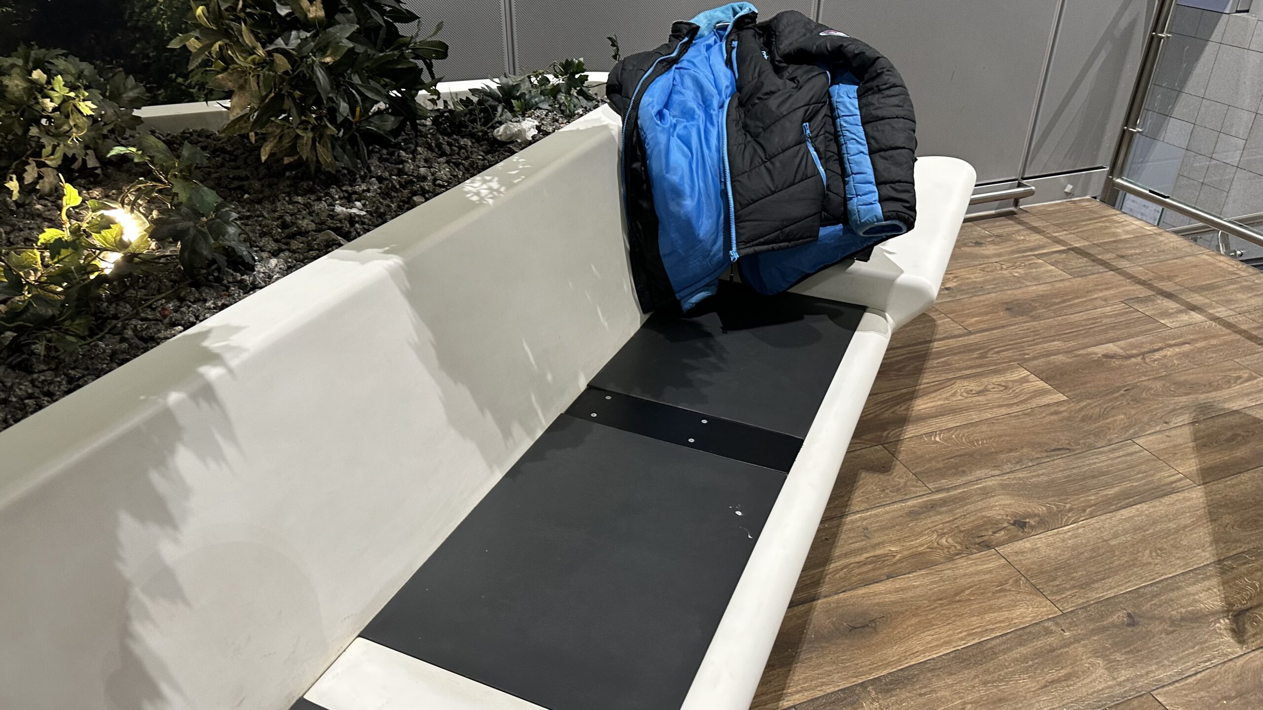 Schlafplatz auf einer weißen Kunststoffbank im Terminal 2 des Frankfurter Flughafens, mit Jacke als Kopfstütze und einer bepflanzten Insel im Hintergrund.
