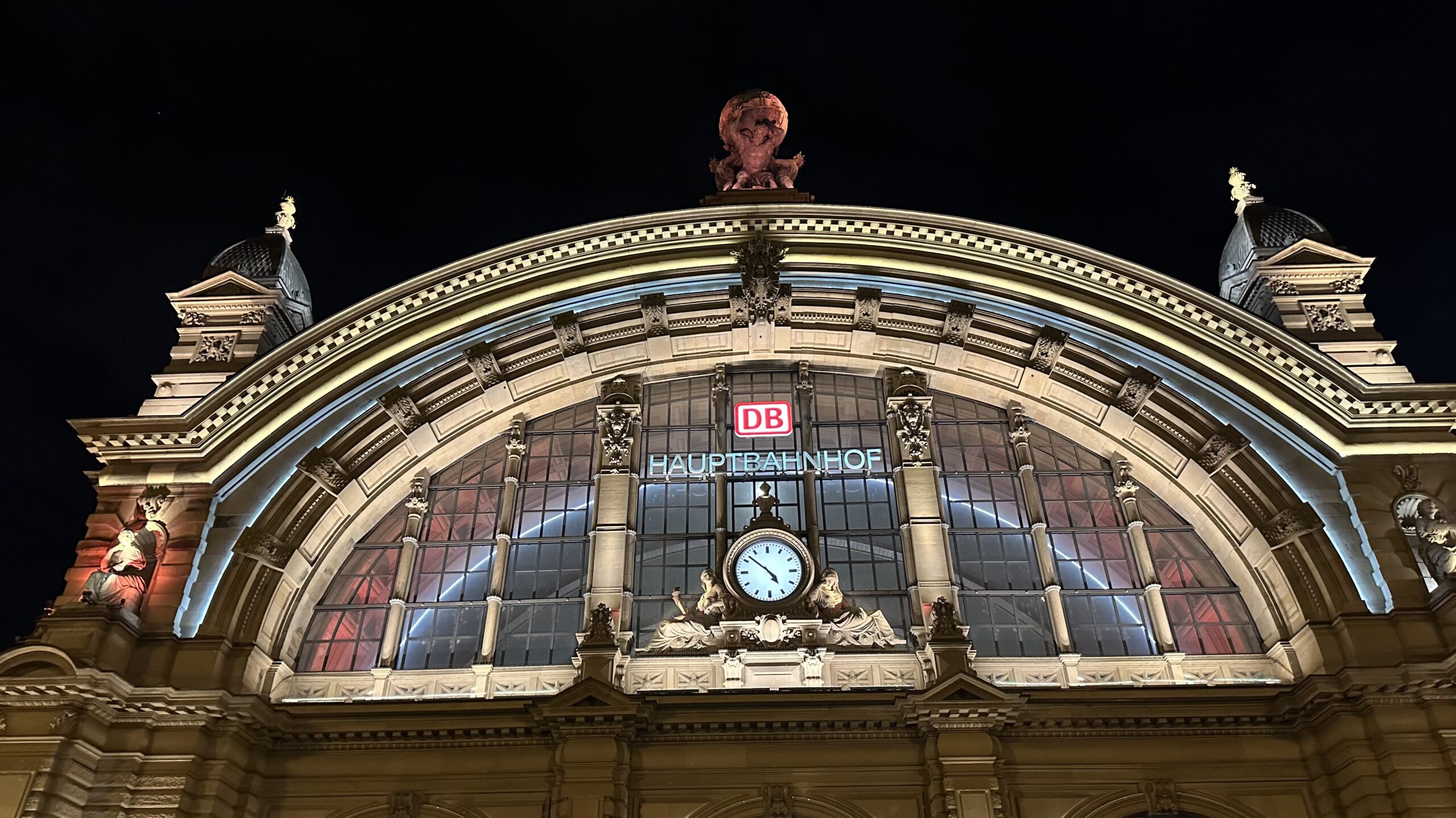 Beleuchtete Fassade des Frankfurter Hauptbahnhofs bei Nacht mit großer Uhr und historischen Figuren über dem Haupteingang.