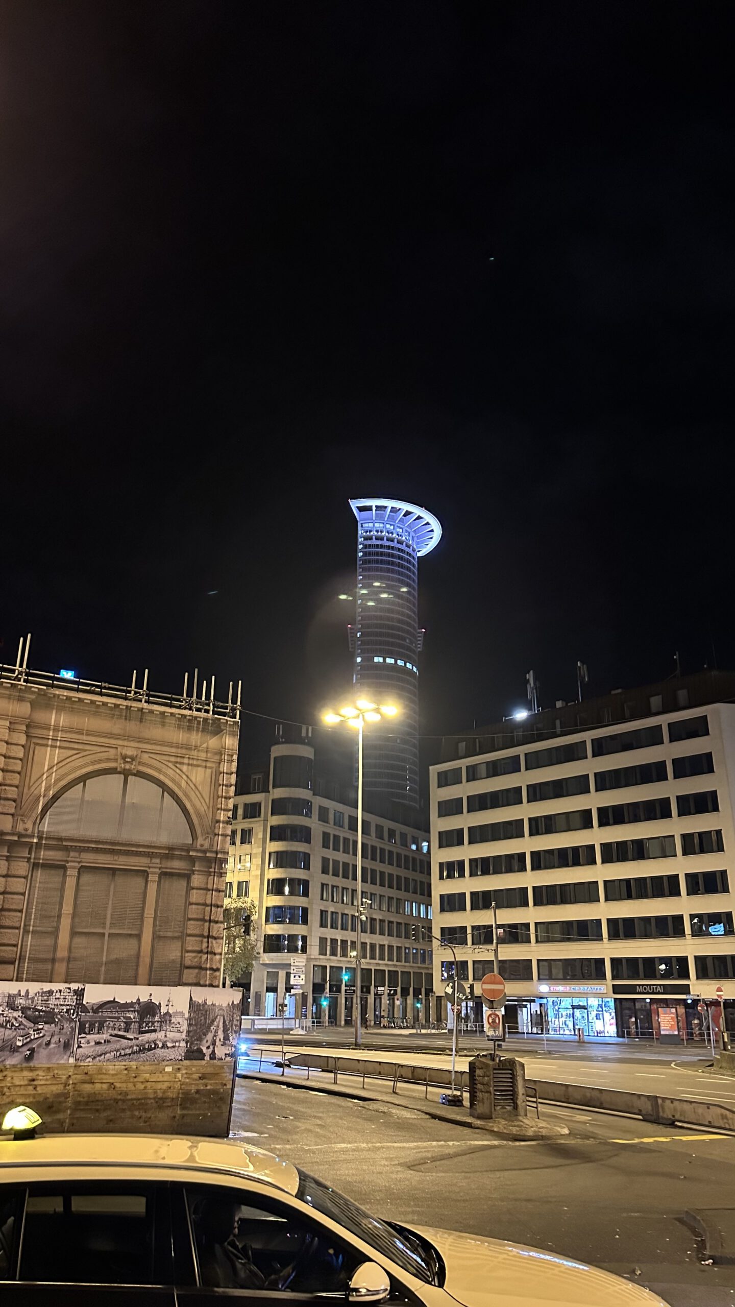 Nachts beleuchtetes DZ-Bank Hochhaus in Frankfurt, gesehen vom Hauptbahnhof aus, mit umliegenden Bürogebäuden und einem Taxi im Vordergrund.