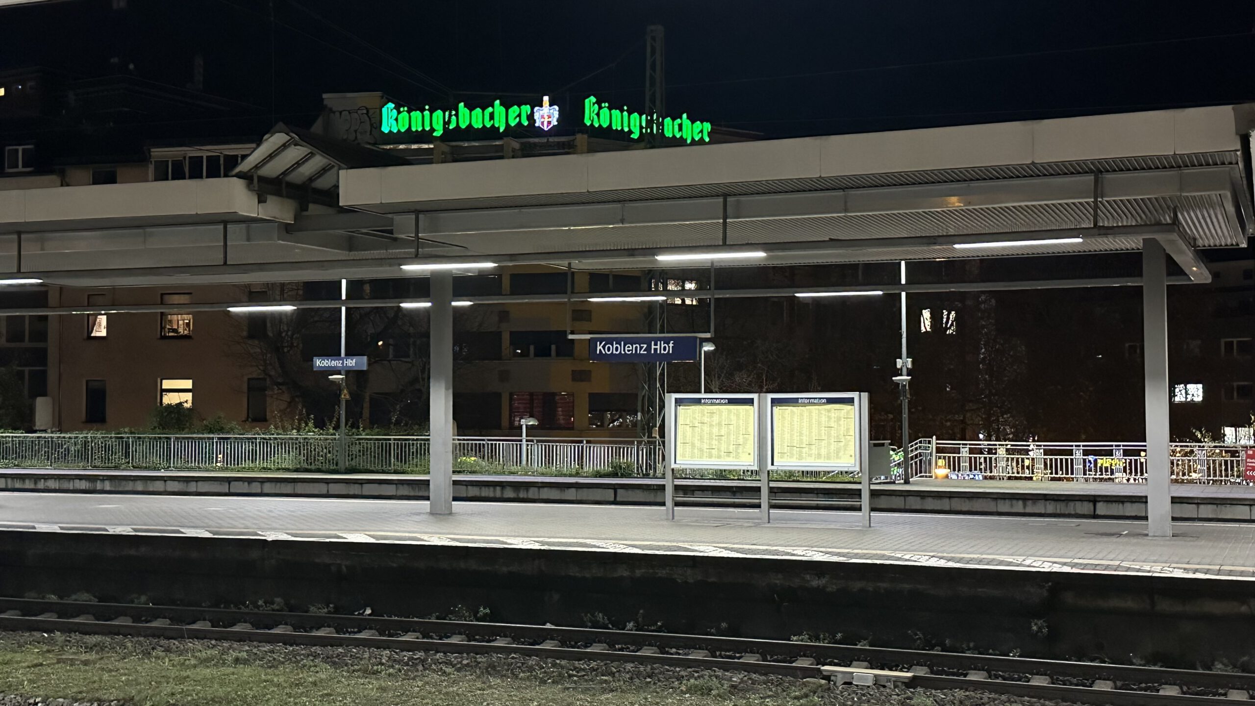 Nachtszene am Hauptbahnhof Koblenz mit beleuchtetem Bahnsteig, Anzeigetafeln und dem grünen „Königsbacher“-Leuchtschild im Hintergrund.