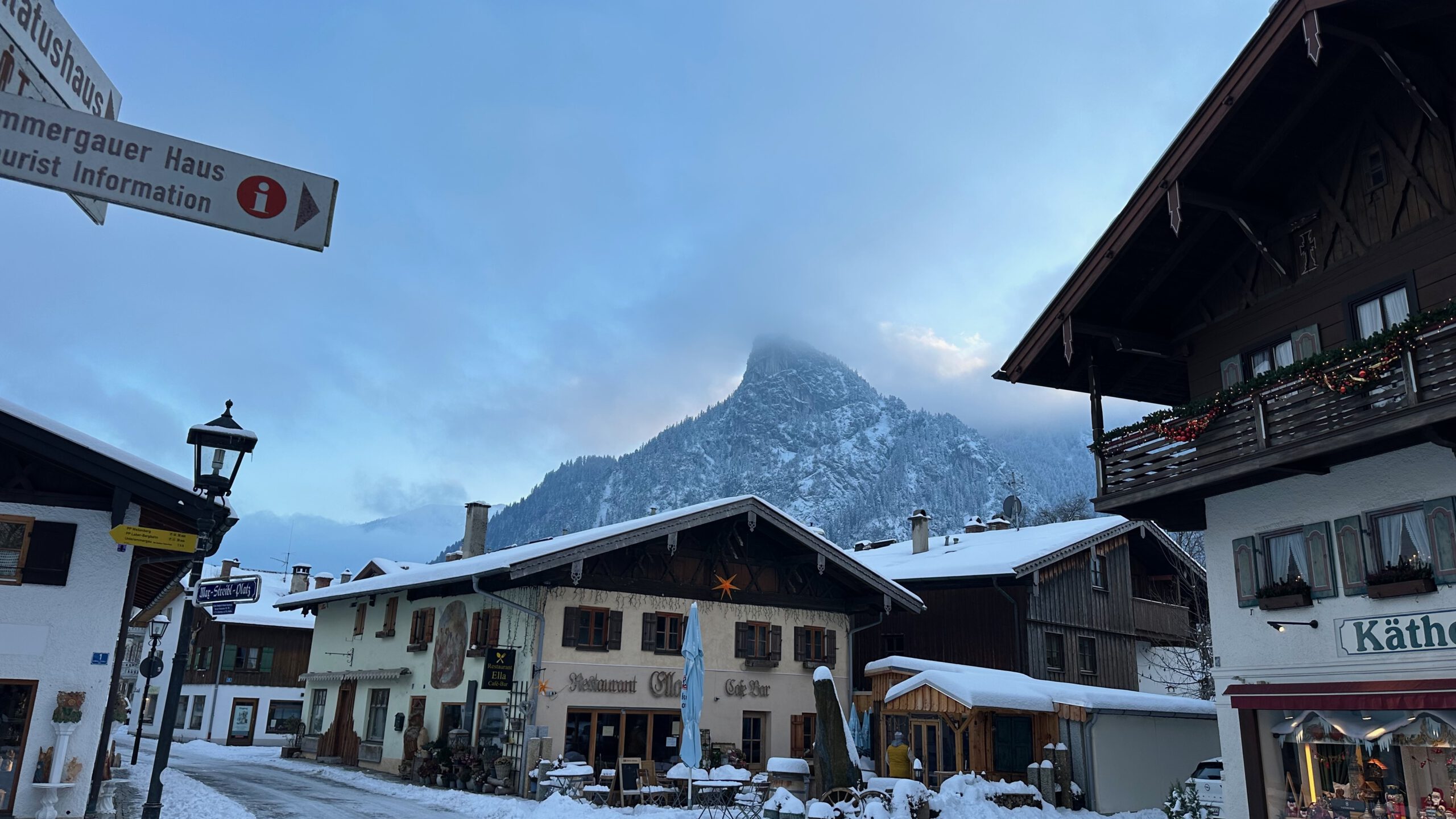 Verschneite Häuser in Oberammergau mit Blick auf den Kofel, dessen Gipfel von dichtem Nebel verhüllt ist.