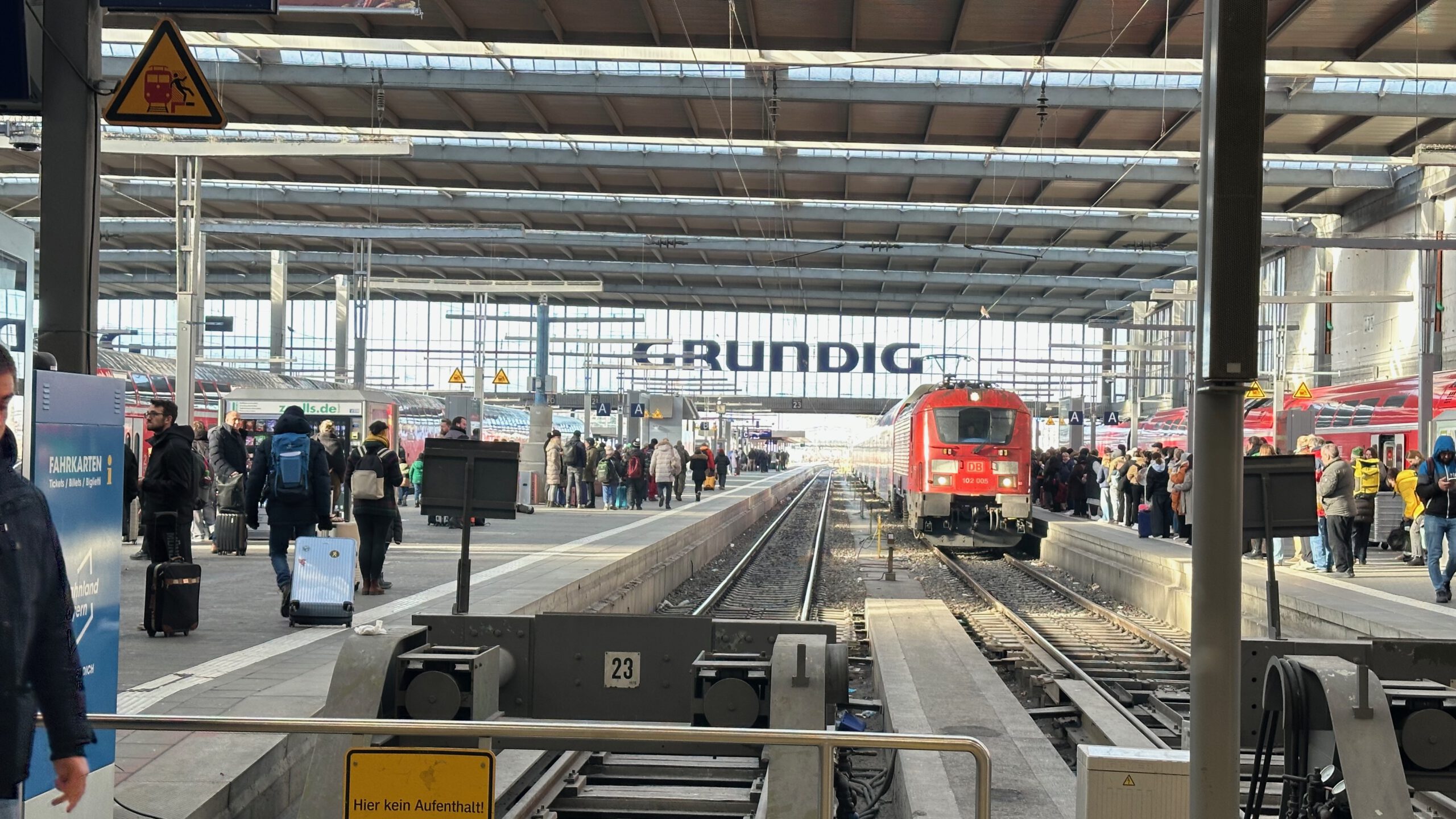 Innenaufnahme des Münchner Hauptbahnhofs mit Gleisen, wartenden Reisenden und einer roten Lok, die am Bahnsteig steht.