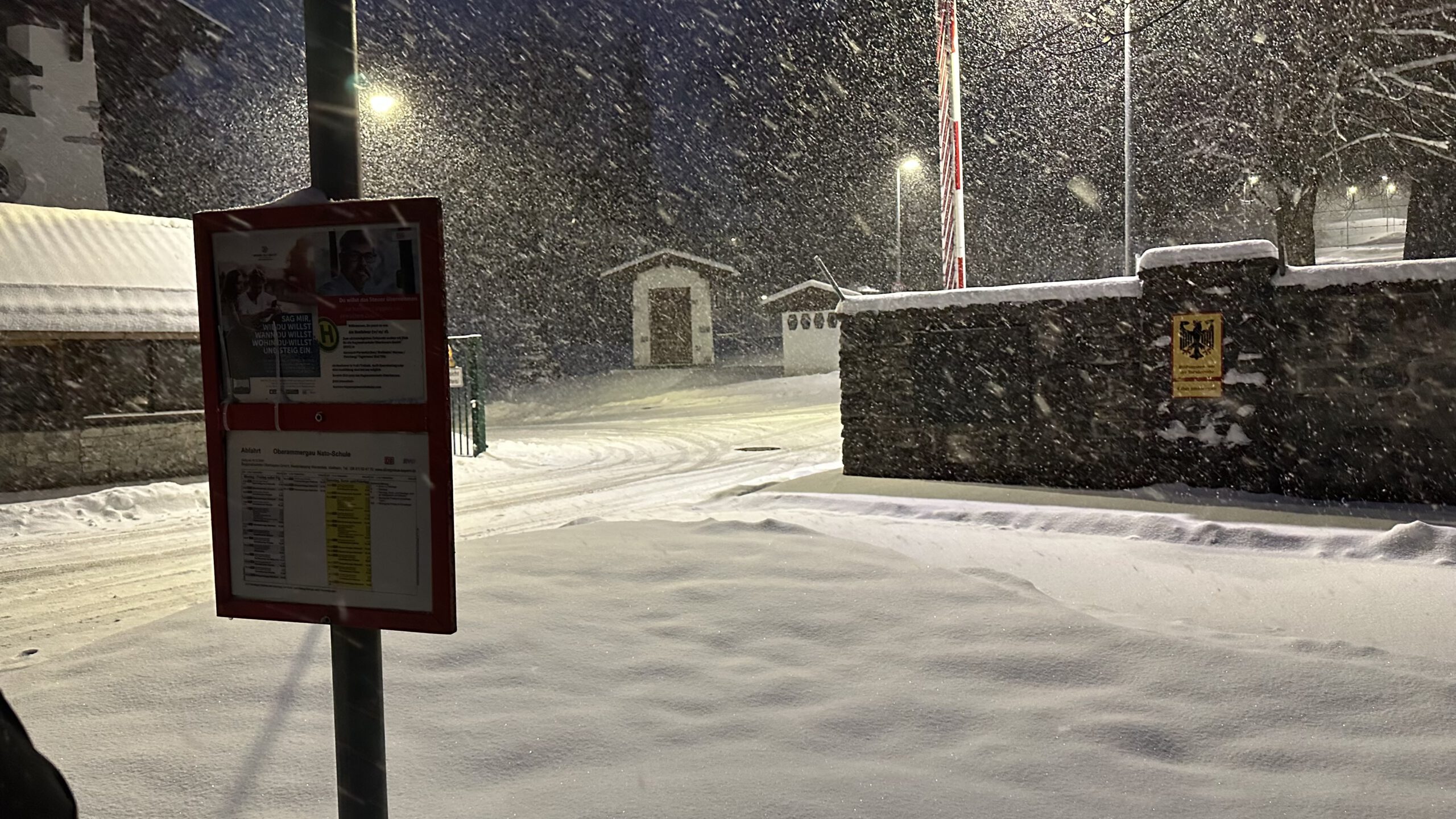 Stark verschneite Einfahrt der NATO School Oberammergau mit Bushaltestelle und Kasernenmauer im dichten Schneefall.