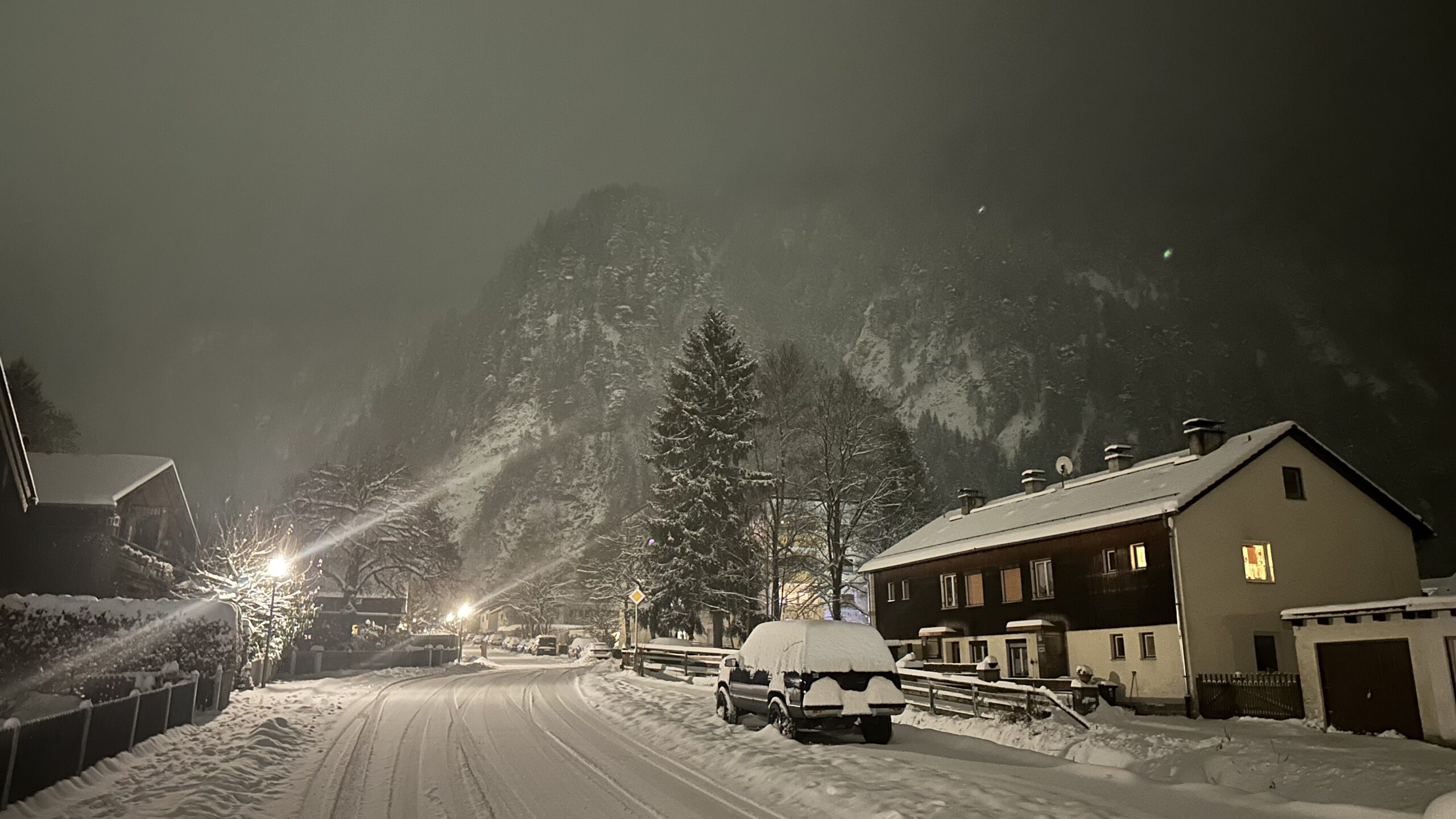 Verschneite Straße in Oberammergau mit beleuchteten Häusern und Blick auf die winterliche Schaffelwand im Hintergrund.
