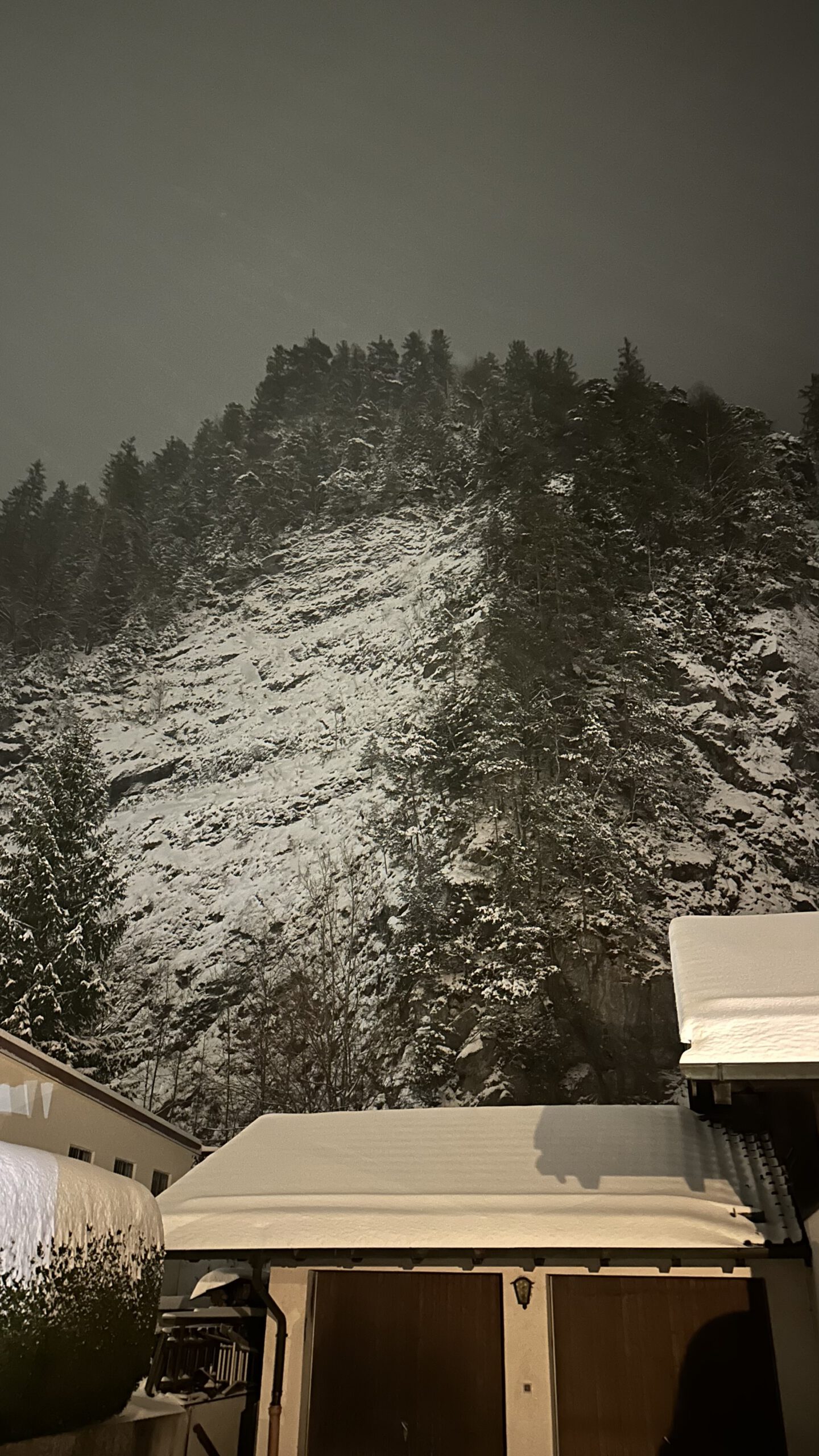 Verschneiter Felshang am Rainenbichl in Oberammergau hinter Wohnhäusern im nächtlichen Schneefall.