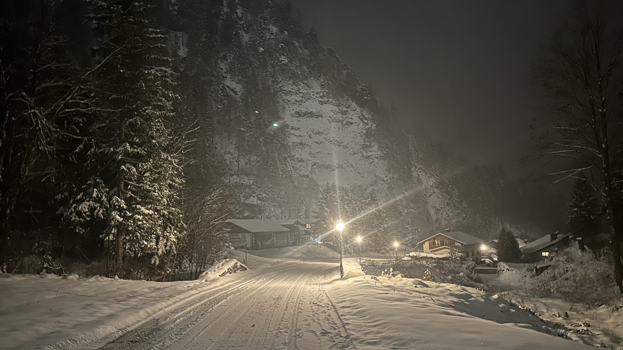 Blick von der NATO School Oberammergau auf die verschneite Schaffelwand bei Nacht, mit beleuchteter Straße, tiefem Schnee und nebelverhangenen Bergen.