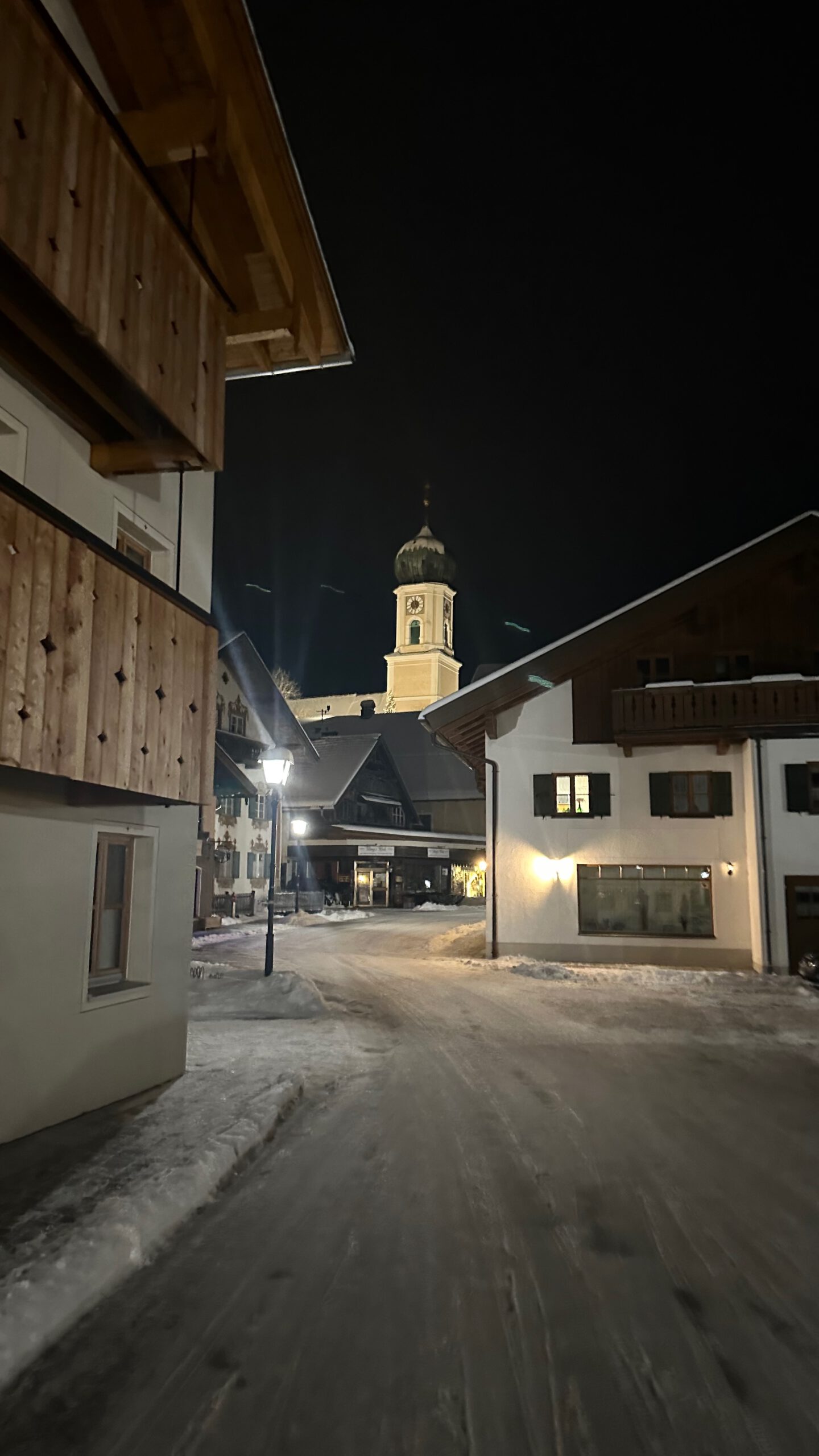 Nächtlicher Blick zur Pfarrkirche Oberammergau Schneebedeckte Gasse in Oberammergau bei Nacht, flankiert von alpenländischen Häusern; im Hintergrund die beleuchtete Pfarrkirche mit Zwiebelturm.