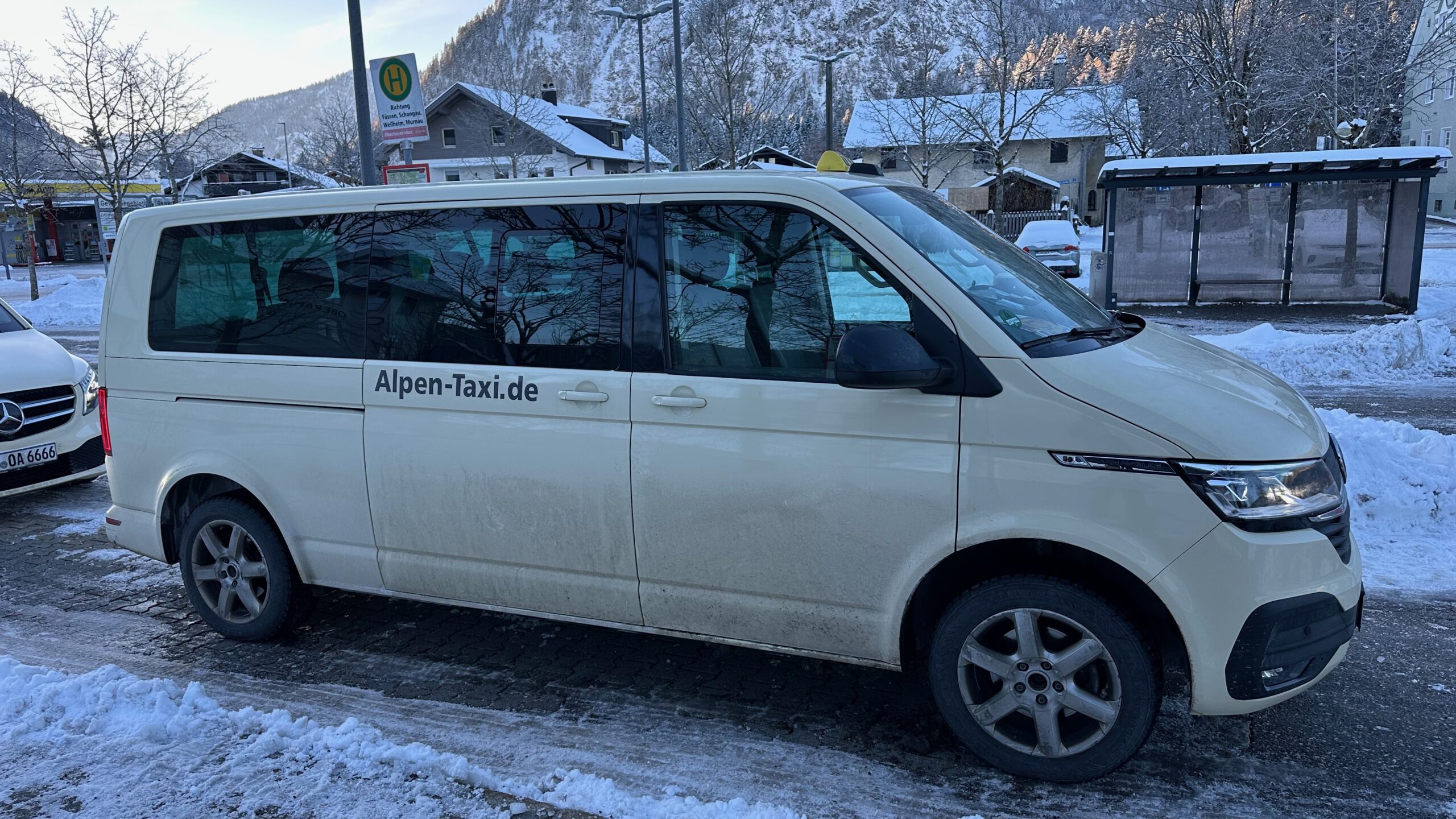 Ein weißer Transporter mit der Aufschrift „Alpen-Taxi.de“ steht im verschneiten Oberammergau und wird als Schienenersatzverkehr genutzt; im Hintergrund Bushaltestelle, Häuser und schneebedeckte Berge.