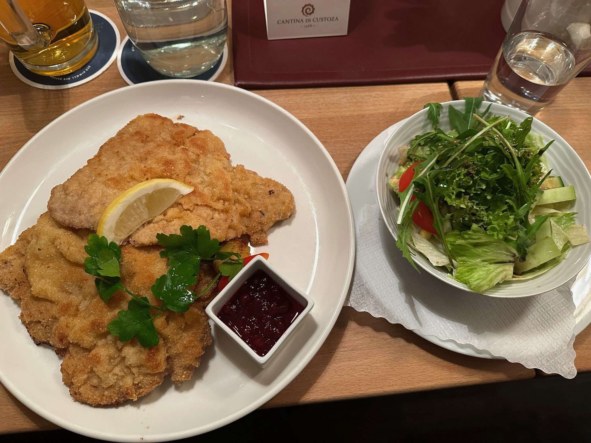 Ein Teller mit zwei goldbraun panierten Wiener Schnitzeln, garniert mit Petersilie und einer Zitronenscheibe, dazu Preiselbeeren und ein Schälchen gemischter Salat daneben.