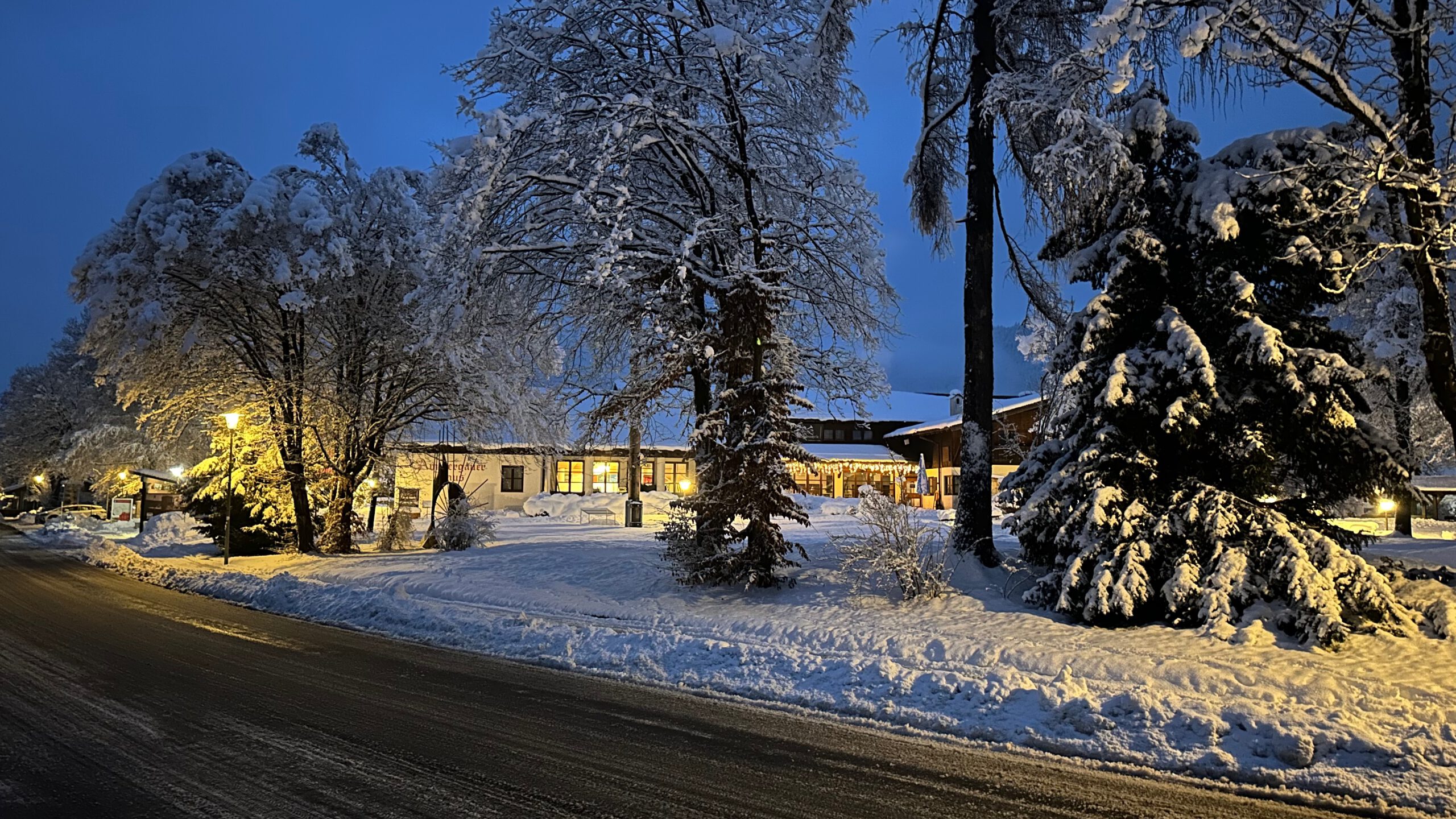 Beleuchtetes Haus am frühen Morgen, umgeben von verschneiten Bäumen und Straßen in Oberammergau.