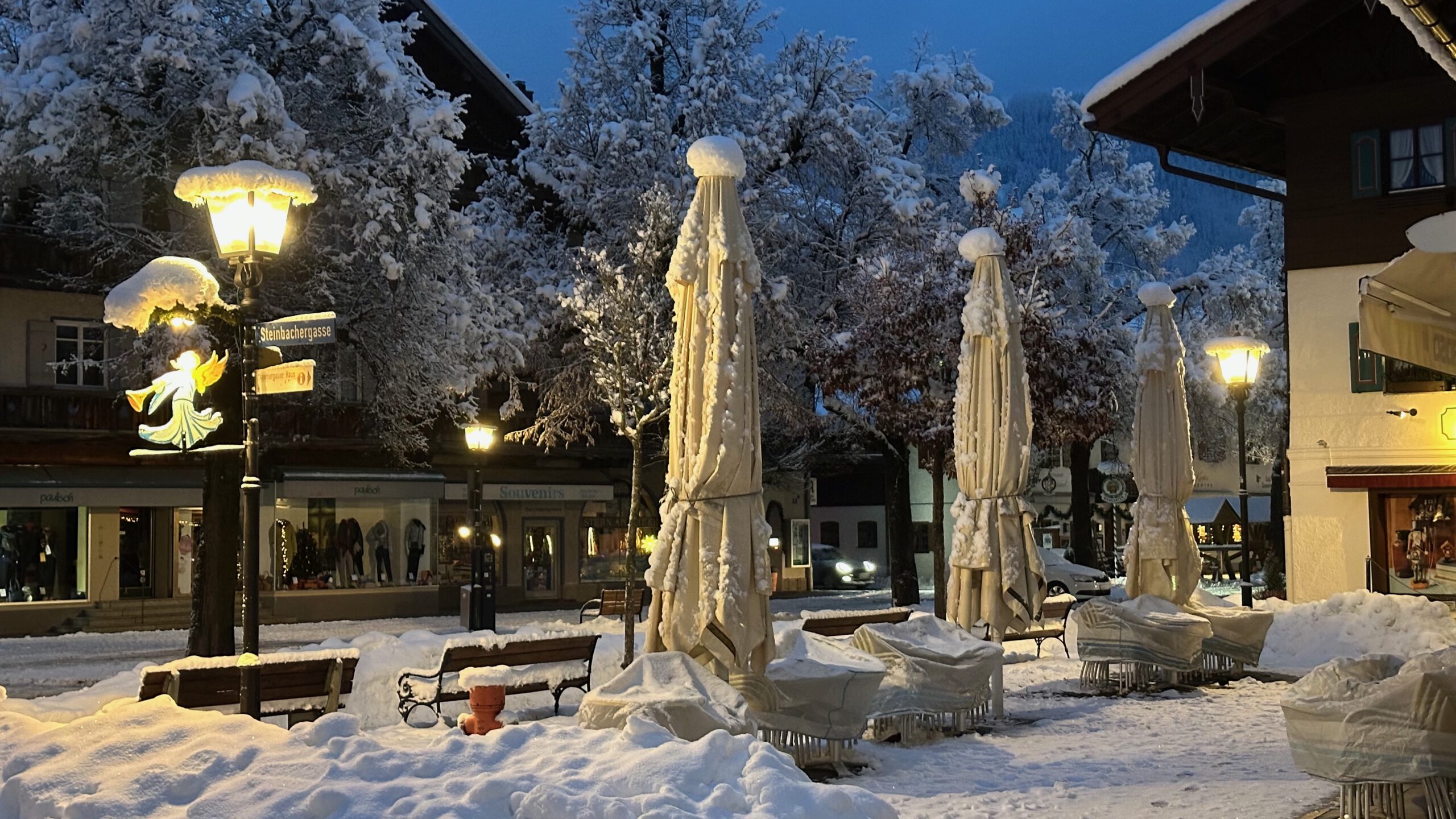 Winterlich verschneite Sonnenschirme und Sitzplätze vor der Eisdiele im Zentrum von Oberammergau.