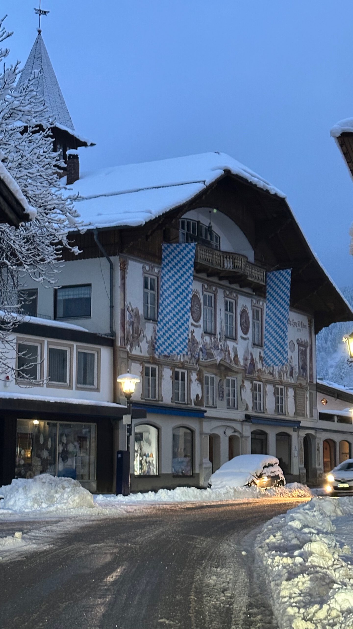 Mit Lüftlmalerei verziertes Haus in Oberammergau mit bayerischen Fahnen und verschneiter Straße am frühen Morgen.