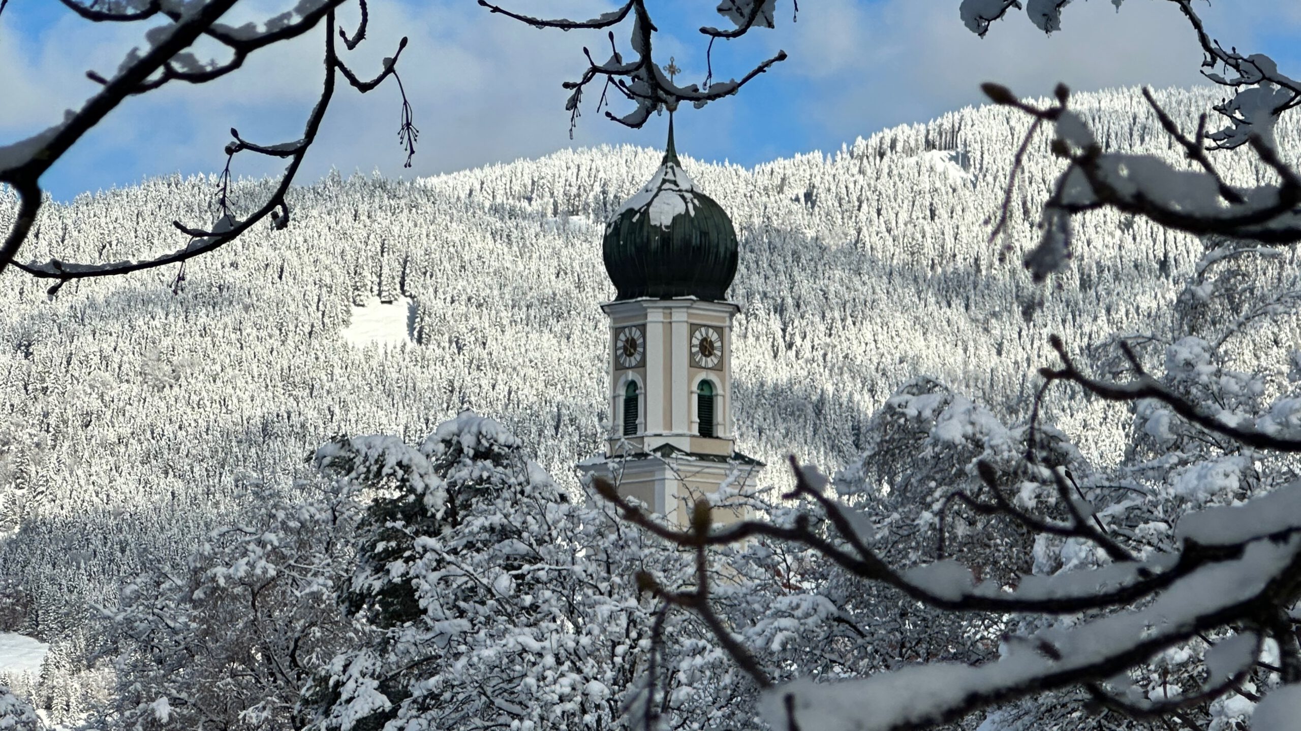 Verschneiter Kirchturm in Oberammergau mit Zwiebeldach, eingerahmt von winterlichen Ästen vor einem schneebedeckten Berghang.