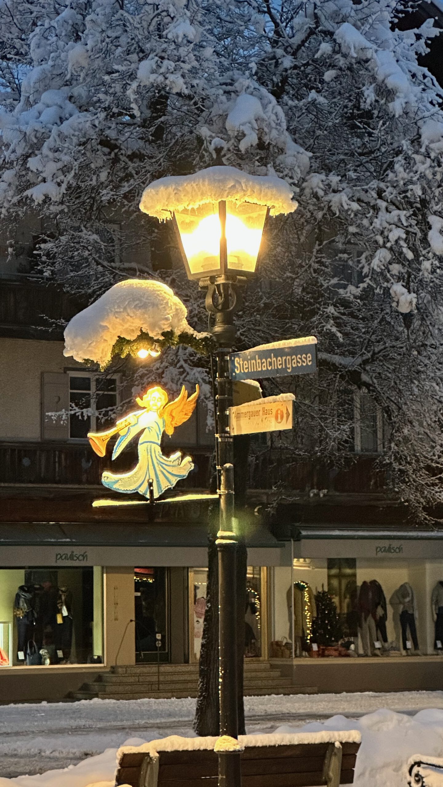Beleuchtete Straßenlaterne in der Steinbachergasse in Oberammergau, mit Schnee bedeckt und mit einer hell leuchtenden Engelsfigur als Weihnachtsdekoration geschmückt.