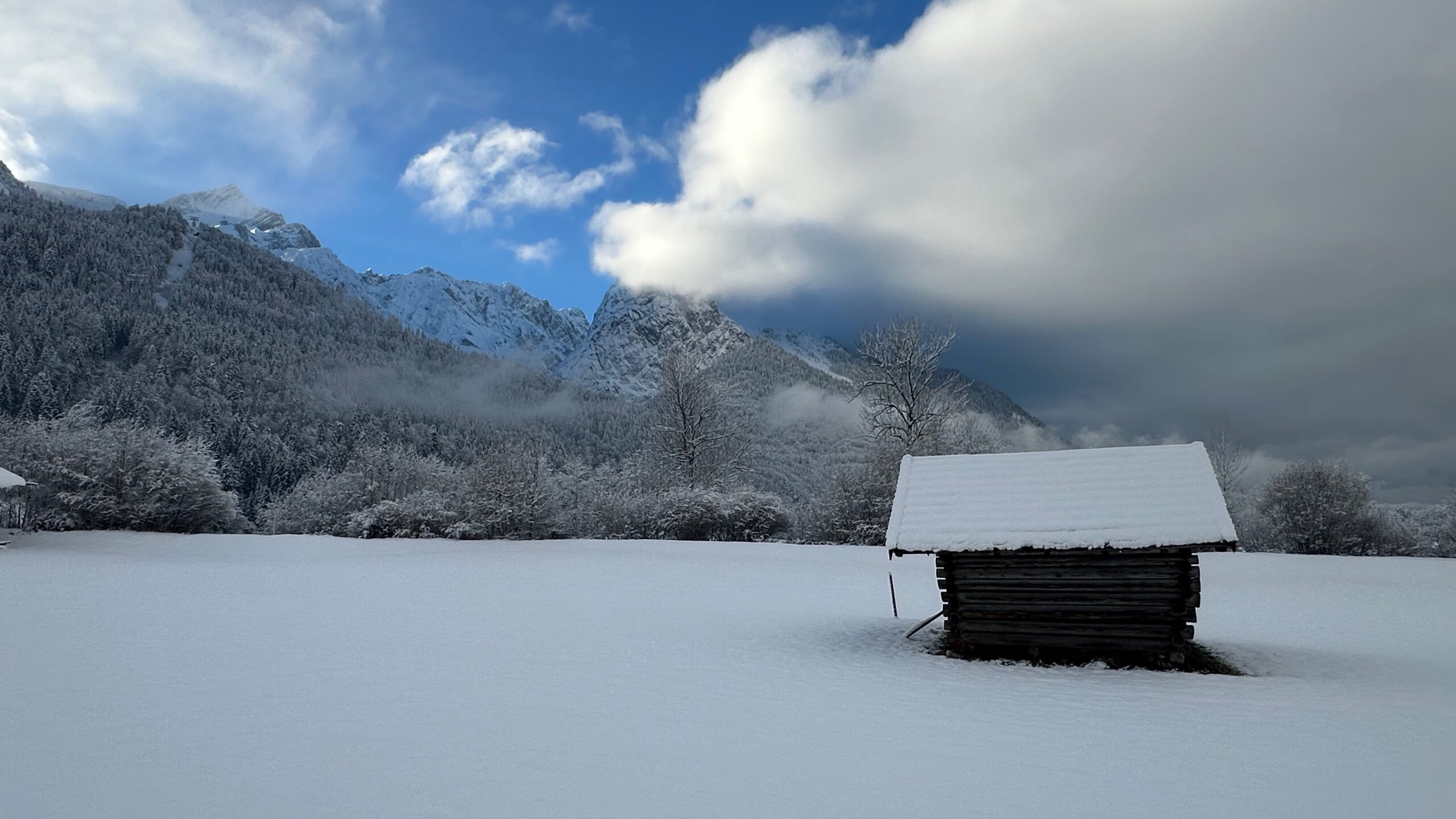 Schneebedeckte Wiese mit einer kleinen Holzhütte im Vordergrund und den bewölkten, winterlichen Bergen bei Garmisch im Hintergrund.