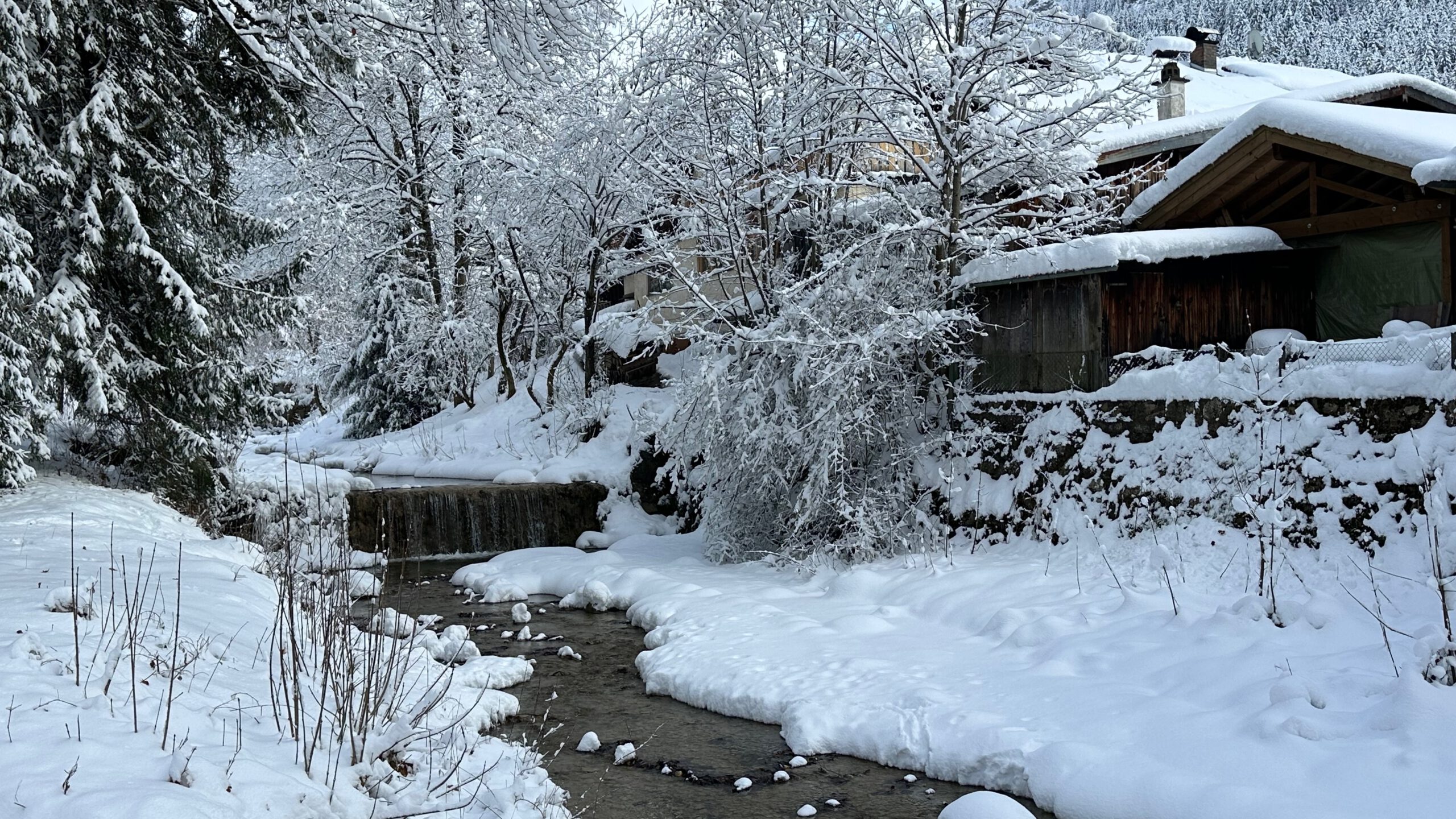 Schmaler Bach in Oberammergau mit kleinem Wasserfall, umgeben von schneebedeckten Bäumen und Häusern im Hintergrund.
