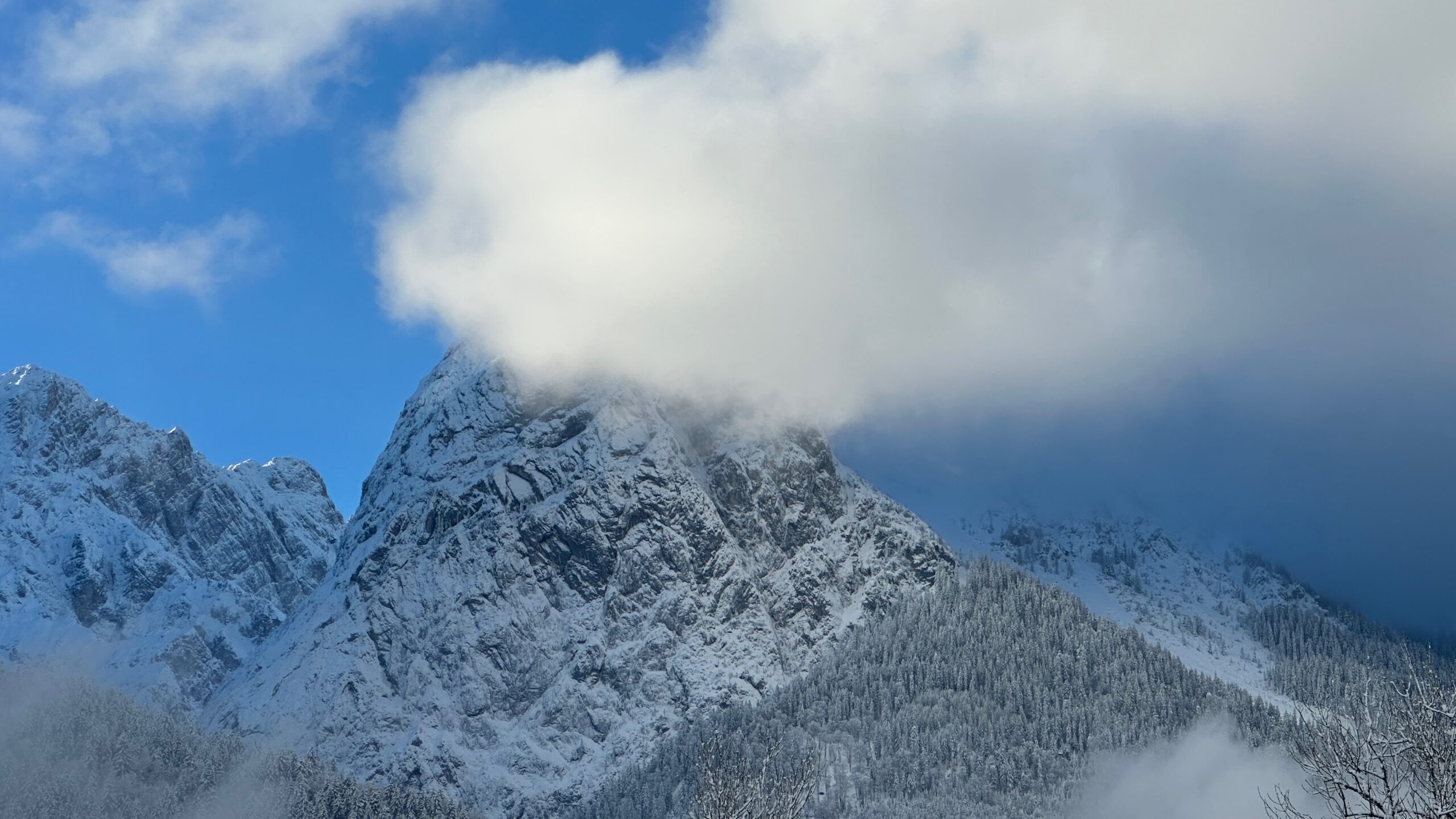 Verschneiter Kofel bei Oberammergau, dessen Gipfel teilweise von tiefhängenden Wolken verdeckt wird.