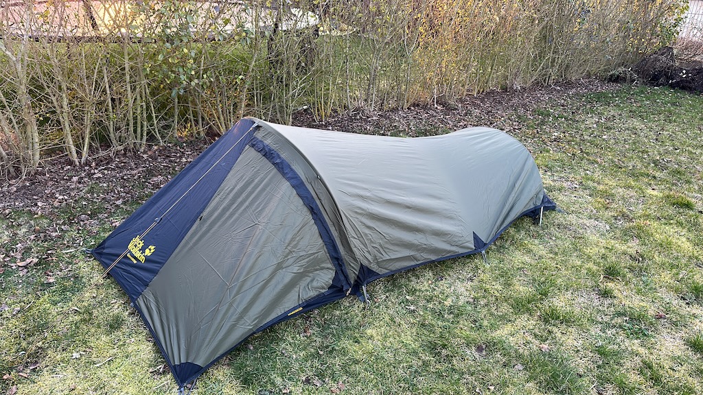 Ein kleines Ein-Mann-Zelt steht aufgebaut in einem Garten auf einer Rasenfläche, umgeben von Hecken – Vorbereitung für eine Nacht im Freien bei kühlen Temperaturen.