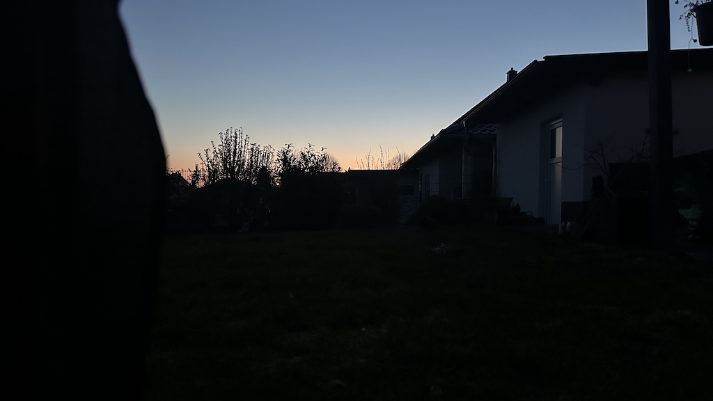 Blick aus einem Zelt am frühen Morgen in einen Garten mit Haus und Hecken, der Himmel zeigt die ersten Lichtfarben der Morgendämmerung.